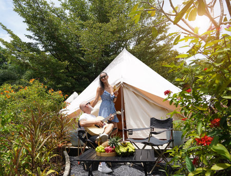 A man plays guitar for a woman near a cozy tent at a glamping site.