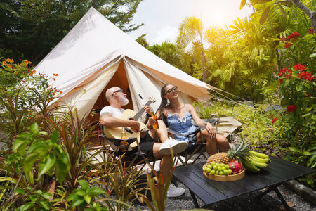 A man and woman is joyfully drinking beer and playing guitar near a cozy tent at glampingの写真素材