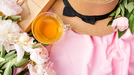 A frame of white and pink peonies, a woman's hat, a cup of tea on a pink fabric background.Floral spring or summer concept.Flat lay. view from above. copyspace. Place for text.の写真素材