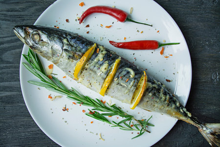 Baked mackerel with lemon and baked potatoes on a white plate. fresh vegetables. Cherry, chili pepper. cutlery. view from above. wooden table. Space for text.の写真素材