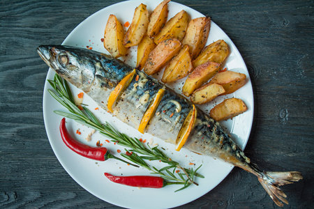Baked mackerel with lemon and baked potatoes on a white plate. fresh vegetables. Cherry, chili pepper. cutlery. view from above. wooden table. Space for text.の写真素材