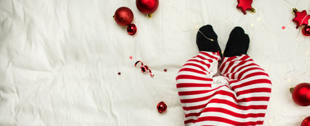 Christmas composition. Legs of a lying newborn with Christmas toys on a white background made of bed linen. Christmas concept. Little Santa Claus. Banner layout, postcard. Flat lay. Copy space.の写真素材