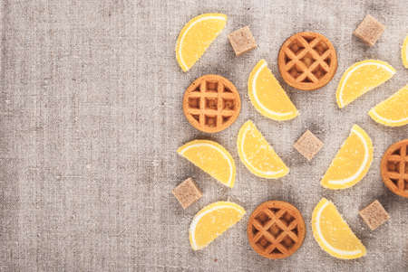 Marmalade pieces in shape of lemon slices, waffles, and brown sugar on rough textile. Sweet food background.の写真素材