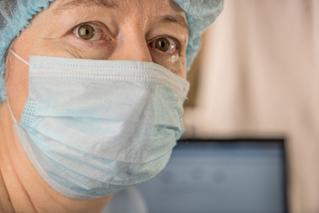 Female doctor wearing medical mask and surgical cap looking seriously and worried at patient with computer in backgroundの写真素材