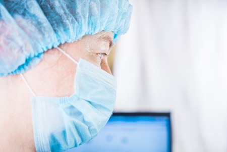 Female doctor wearing medical mask and surgical cap looking seriously and worried at patient with computer in backgroundの写真素材