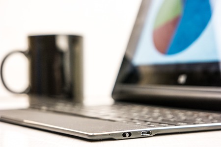 Black laptop with business charts on screen, on white table in office. With coffe cup in background.の写真素材