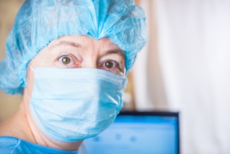 Female doctor wearing medical mask and surgical cap looking seriously and worried at patient with computer in backgroundの写真素材