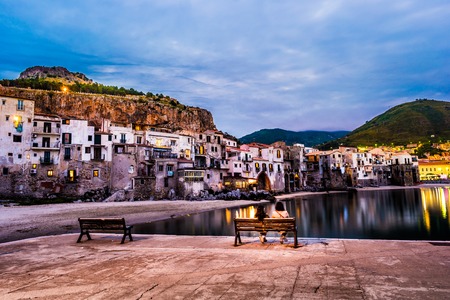 Unrecognizable couple on a bench with view on habour and old houses in Cefalu at night, Sicily. Beautiful townscape of old italian town. Travel photography.の写真素材