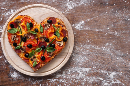 Baked heart-shaped homemade pizza on a cutting board on rustic table, close-up.