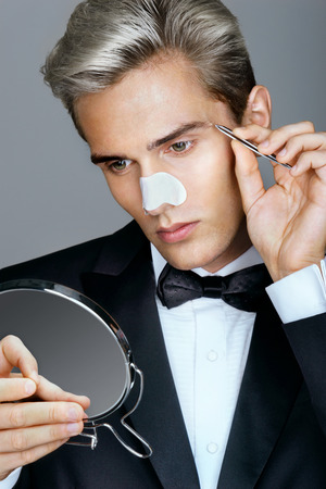 Young man tweezing the hair on his face. Photo of gentleman looking in the mirror, cares for his appearance. Grooming himselfの写真素材