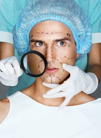 Nurse with magnifying glass near the face of the patient. Close up of male with pencil marks on skin for cosmetic medical procedures. Surgical mark lines on eyes, nose, cheek, and jawの写真素材