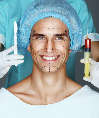 Nurse with syringe and scalpel near the face of scared patient. Happy man with pencil surgical lines (forehead, eyes, nose, cheekbone and jaw). Plastic Surgery conceptの写真素材