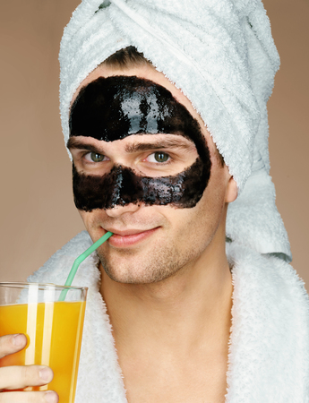 Well groomed man with black mask on the face, drinks orange juice. Photo of smiling man receiving spa treatments. Healthy and happy conceptの写真素材