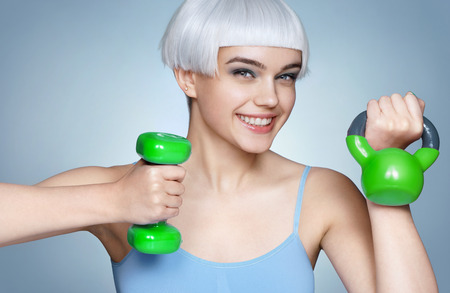 Happy young girl with green dumbbell and kettlebell in her hands. Photo of fashion blonde girl on blue background. Close up. Healthy lifestyle conceptの写真素材
