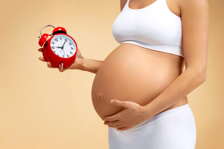 Close up of pregnant woman holding alarm clock on beige background. Pregnancy, maternity,...
