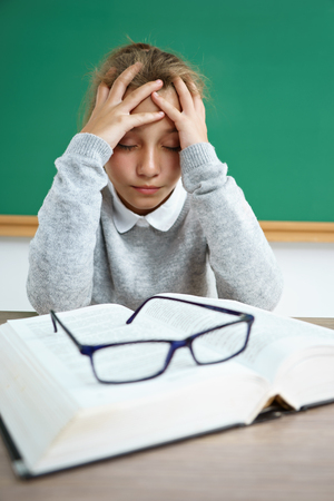 Upset girl holding her head. Photo of schoolgirl with book on the desk in classroom. Back to schoolの写真素材