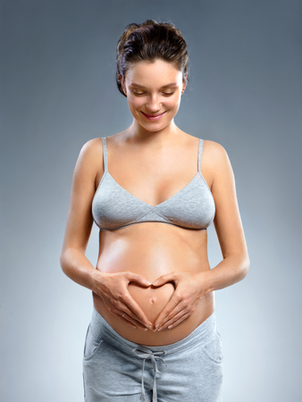 Smiling pregnant woman making a heart gesture with her fingers on grey background. Pregnancy, maternity, preparation and expectation conceptの写真素材