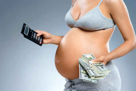 Close up of pregnant woman holding cash money and calculator on grey background. Pregnancy, maternity, preparation and expectation conceptの写真素材