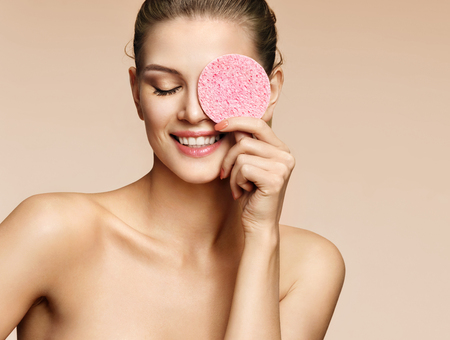 Smiling woman with cosmetic sponge covering one eye. Photo of woman with perfect skin on beige background. Beauty conceptの写真素材