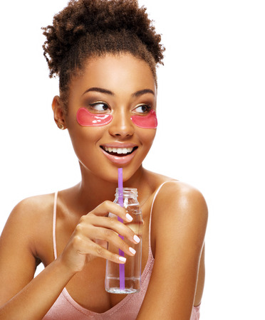 Beautiful girl with pink eye patches on her face and holds bottle of clean water. Photo of young african american girl isolated on white background. Youth and Beautyの写真素材