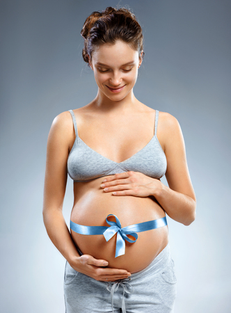 Pregnant woman with blue ribbon and touching her belly on grey background. Pregnancy, maternity, preparation and expectation conceptの写真素材