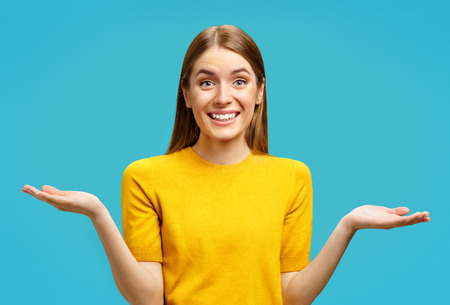 Smiling young girl looks at camera and spreads hands. Photo of attractive girl shows open palms with something in yellow sweater on blue backgroundの写真素材
