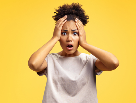Surprised girl holds hands on head, stares in terror. Photo of african american girl wears casual outfit on yellow background. Emotions and omg conceptの写真素材