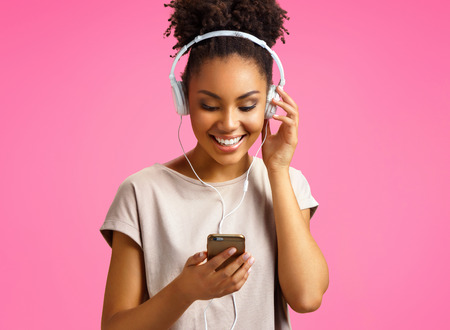 Happy young girl listens music in headphones. Photo of african american girl wears casual outfit on pink background. Emotions and pleasant feelings concept.の写真素材