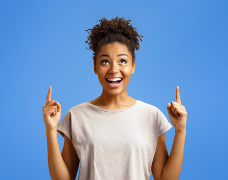 Amazed girl looks up, indicates with two forefingers upwards at copy space, shows something. Photo of african american girl on blue background. Emotions and pleasant feelings concept.の写真素材