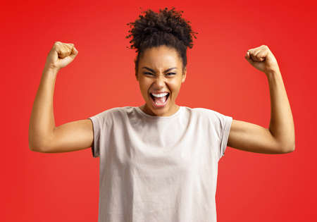 Overjoyed girl clenches fists in triumph. Photo of african american girl wears casual outfit on red background. Emotions and pleasant feelings concept.の写真素材