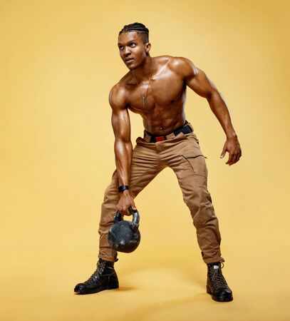Young muscular guy training with kettlebell. Photo of handsome man with naked torso on yellow background. Strength and motivationの写真素材