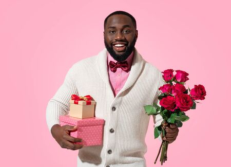 Handsome man holds gifts and bouquet of roses. Photo of african american man expresses love to someone, romantic feelings on pink background. Be my Valentineの写真素材