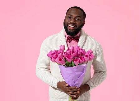 Handsome man holds bouquet of spring flowers. Photo of african american man expresses love to someone, romantic feelings on pink backgroundの写真素材