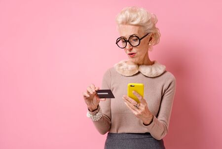 Problem with a credit card. Grandmother makes a payment, using a credit card and smartphone. Photo of kind elderly woman on pink background.の写真素材