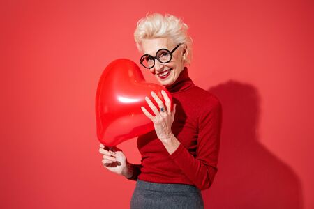 Lovely woman hugs red heart shape balloon. Photo of smiling elderly woman in love on red background. Valentine's Dayの写真素材