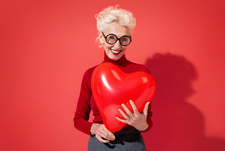 Lovely woman hugs red heart shape balloon. Photo of smiling elderly woman in love on red background....