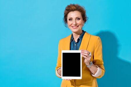 Smiling elderly woman holds digital tablet. Photo of business lady in yellow blazer on blue background. Emotions and pleasant feelings concept.の写真素材