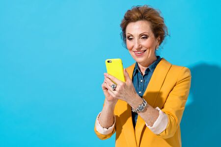 Smiling elderly woman using phone. Photo of business lady in yellow blazer on blue background. Emotions and pleasant feelings concept.の写真素材