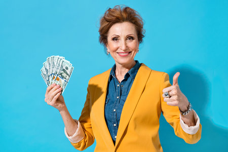 Happy elderly woman holds cash money and like gesture. Photo of business lady in yellow blazer on...