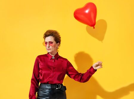 Frustrated woman because feelings of love left her, with a flying away red heart-shaped balloon as a symbol of parting with love. Photo of elderly woman in love in red shirt on yellow backgroundの写真素材