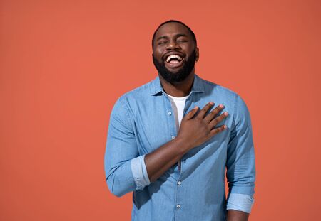 Overjoyed man holding his hands on heart can't stopping laughing. Photo of african man in casual outfit on coral background.の写真素材