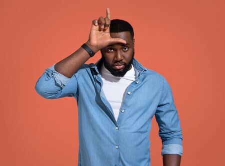 Man making fun of people with fingers on forehead doing loser gesture mocking and insulting. Photo of african man in casual outfit on coral background.の写真素材