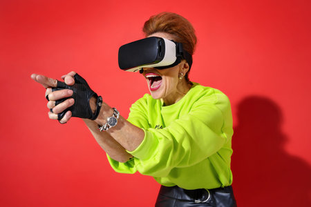 Forever Young! Elderly woman in bright outfit in VR glasses shooting on red backgroundの写真素材