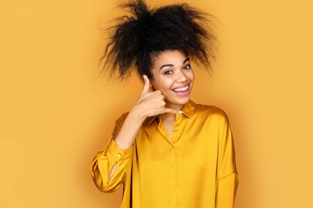 Smiling girl makes phone gesture of calling, call me back sign. Photo of african american girl on yellow backgroundの写真素材