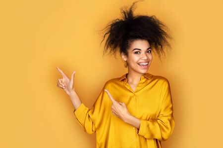 Smiling girl points away with forefingers, shows on copy space for your text. Photo of african american girl on yellow backgroundの写真素材