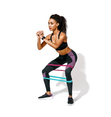 Sporty woman doing squats with resistance band. Photo of african american girl in black sportswear on white background. Strength and motivationの写真素材