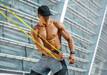 Athletic man performs exercises using resistance band. Photo of strong man training in the city....