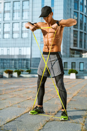 Muscular man performs exercises using a resistance band. Photo of young man training in the city....