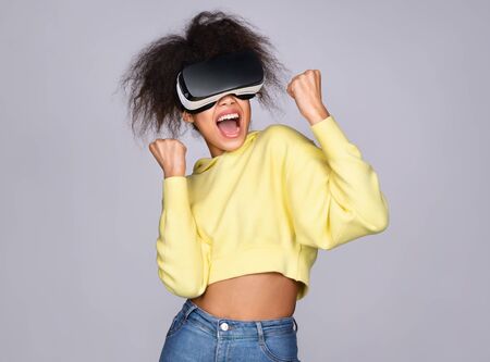 Happy girl holding fists up and celebrating virtual win wearing VR-glasses on grey backgroundの写真素材