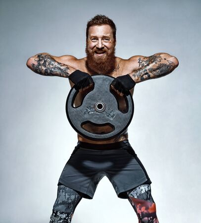Athletic man doing exercise with heavy weights plate. Photo of man with naked torso on grey background. Strength and motivationの写真素材